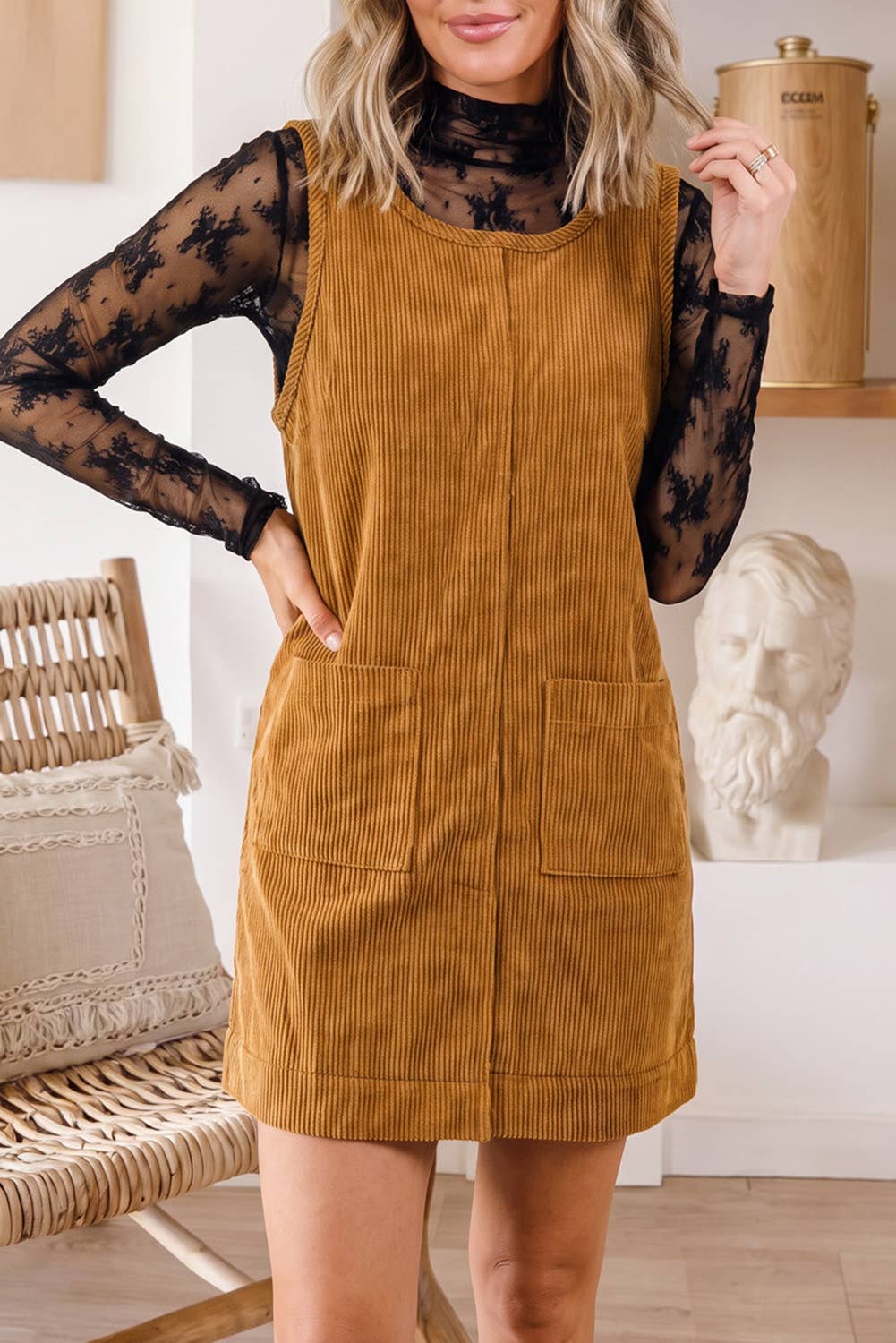 A woman models a mustard yellow corduroy pinafore dress over a black sheer long-sleeve t-shirt in a bright, modern room setting.