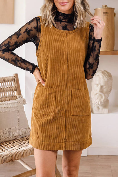 A woman models a mustard yellow corduroy pinafore dress over a black sheer long-sleeve t-shirt in a bright, modern room setting.