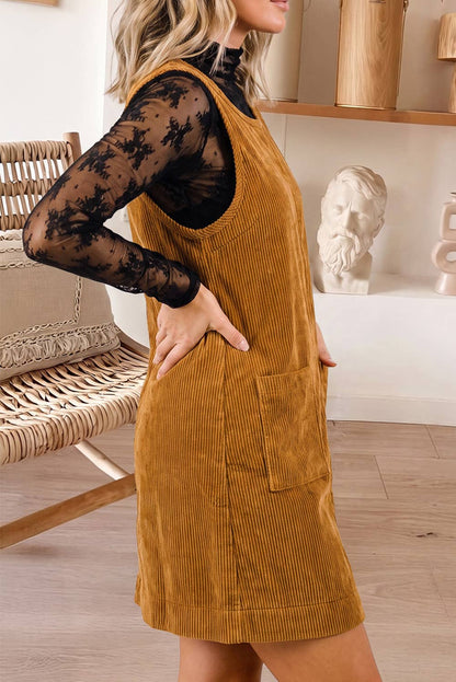 A woman wearing a mustard yellow corduroy pinafore dress over a black lace long-sleeve top, standing in a bright room with a wicker chair and shelf decor in the background.