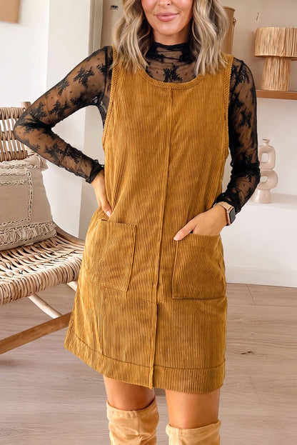 A woman wearing a sleeveless mustard yellow corduroy pinafore dress over a black sheer long-sleeve top, paired with tan knee-high boots, in a brightly lit indoor room.